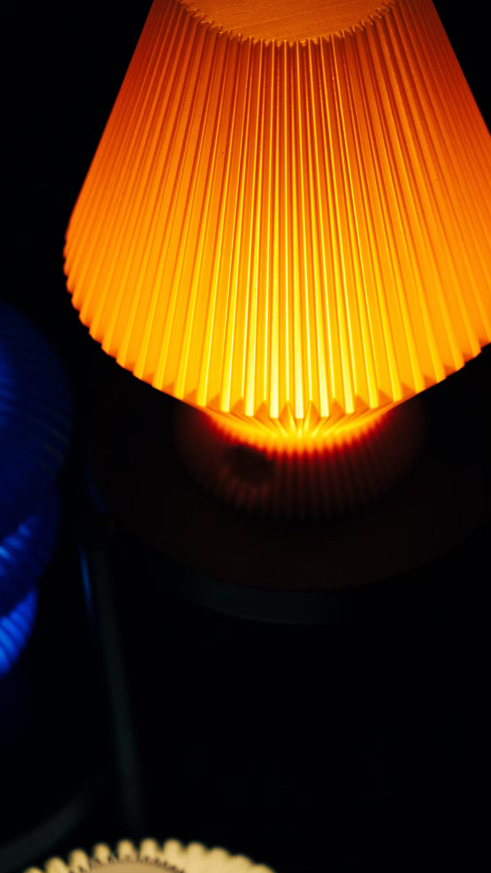 Close-up of an orange Muvèlo sculptural portable table lamp glowing warmly against a dark background.