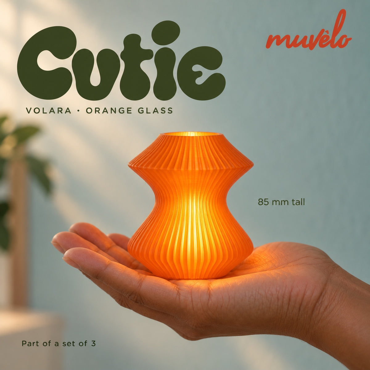 Illuminated Orange Glass Volara Cutie resting on an adult palm, with soft retro Cutie lettering and the official Muvèlo wordmark.