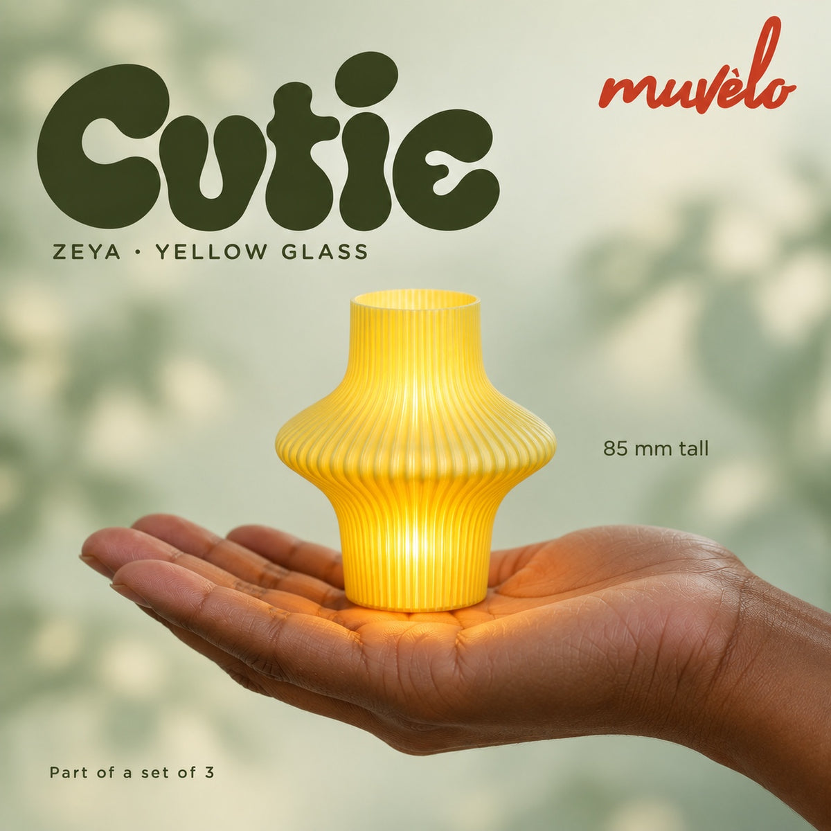 Illuminated Yellow Glass Zeya Cutie resting on an adult palm, with soft retro Cutie lettering and the official Muvèlo wordmark.