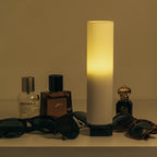 Lamp with ribbed design emitting a warm glow, surrounded by sunglasses and bottles on a beige surface.