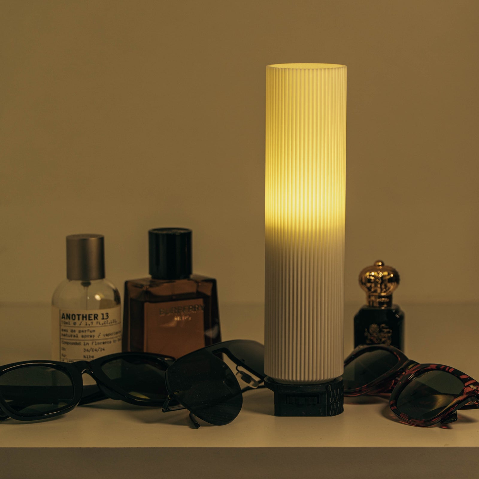 Lamp with ribbed design emitting a warm glow, surrounded by sunglasses and bottles on a beige surface.