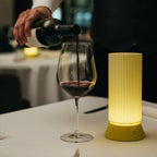 Person pouring wine into a glass on a table with a lamp and cutlery.