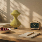 Yellow vase, bowl of apples, alarm clock, and notebook on a wooden surface with a warm light.